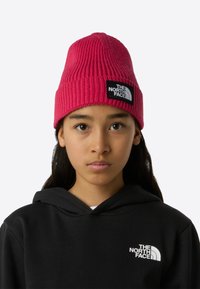 Vaaleanpunainen neulottu pipo, jossa on ribbineulos ja logopläntti. Sen käyttää henkilö, jolla on musta huppari, jossa on kengurun tasku ja kiristysnauhallinen huppu.