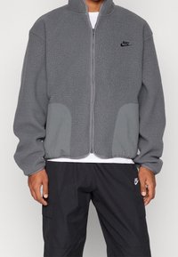 Giacca in pile grigia con colletto alto, chiusura con zip e tasche grigie a contrasto. Presenta il logo Nike in modo discreto sul petto. Indossata sopra pantaloni neri.