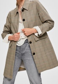 Rutet frakke i beige, brune og grå nuancer. Lavet af struktureret stof med krave, knaplukning og sidelommer. Bæres over en stribet bluse.
