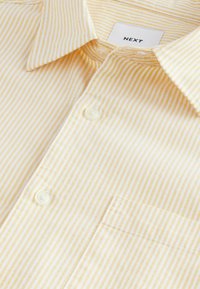 Yellow and white striped shirt with a button-down collar, featuring a single chest pocket and a tag labeled "NEXT". Soft fabric texture.