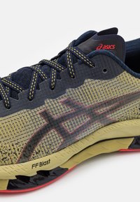 La chaussure de course Asics présente un dessus en maille jaune respirant avec des accents bleu foncé, un design à motifs et une semelle en caoutchouc noire et rouge.