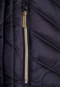 Sección de chaqueta acolchada azul marino con acolchado diagonal, que presenta un destacado zipper de metal dorado y un tirador de zipper. Textura de tela lisa y brillante.