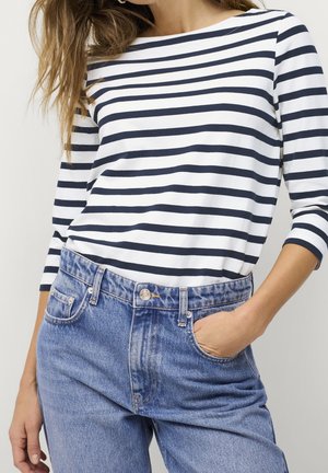 Femme portant un haut à manches longues rayé horizontal noir et blanc, rentré dans un jean en denim bleu taille haute, une main dans la poche.