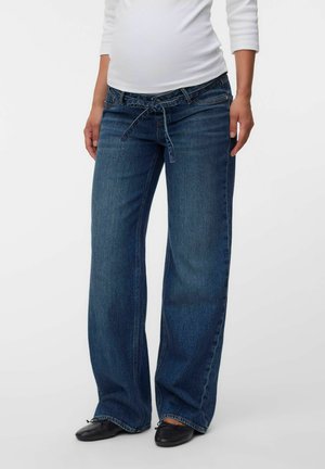 Femme portant un jean bleu ample avec une taille nouée et des chaussures plates noires, debout devant un fond uni.
