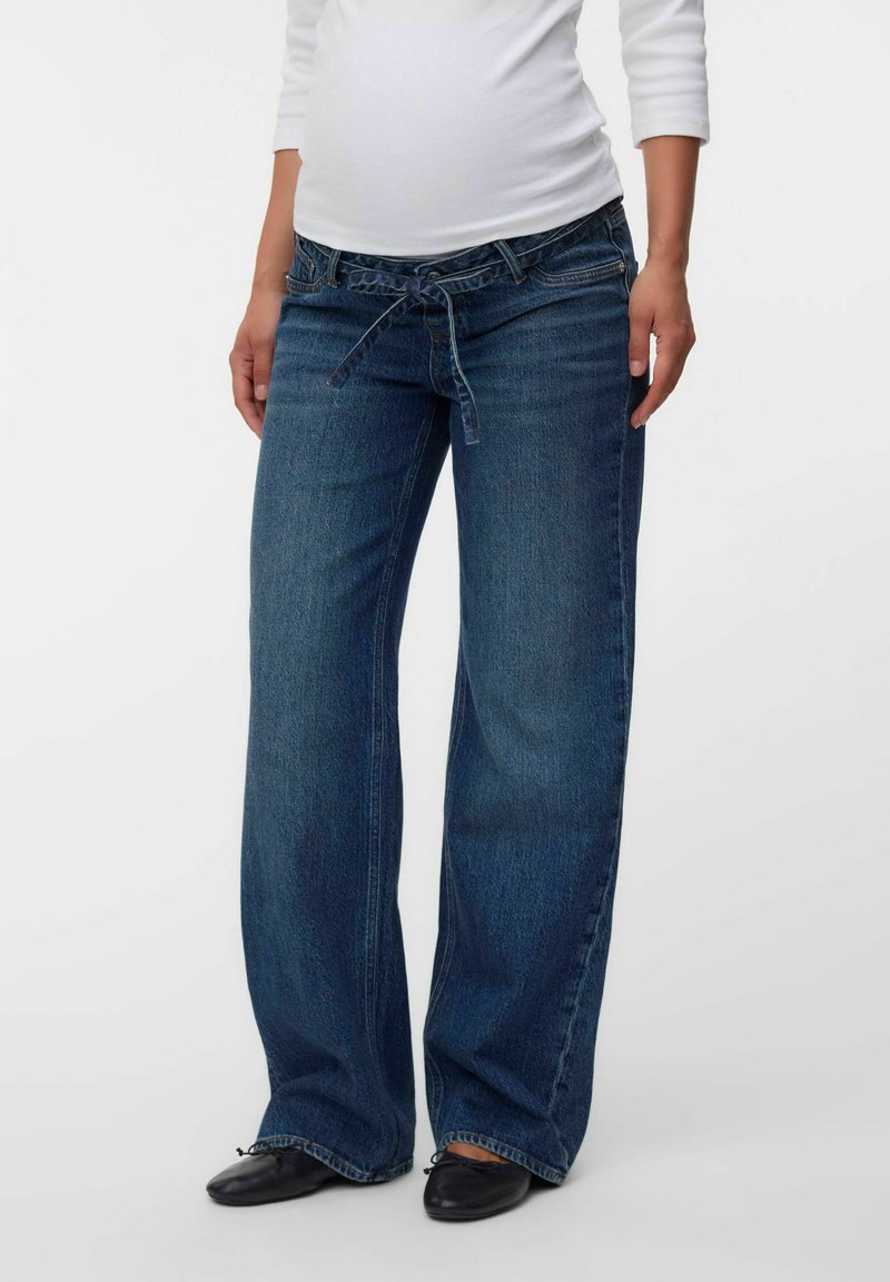 Femme portant un jean bleu ample avec une taille nouée et des chaussures plates noires, debout devant un fond uni.