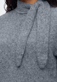Gros plan d'une personne portant un pull gris doux et texturé avec un col montant et des liens larges attachés ressemblant à une écharpe.