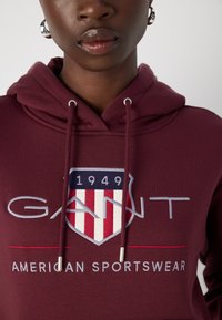 Felpa bordeaux con grande logo scudetto ricamato, strisce rosse e bianche, hardware argentato e cordini. "GANT" in testo bianco.