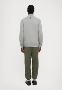 Mann iført grå langermet topp med svarte striper på øvre rygg, olivengrønne mønstrede bukser, hvite sokker og svarte sneakers, står vendt bort.
