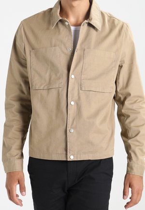 Man wearing beige button-up jacket with two chest pockets and black pants, standing with arms relaxed at sides.