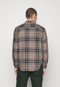 Ruskea ruudullinen paita, jossa sinisiä ja vihreitä yksityiskohtia, napilliset kalvosimet ja kaareva helma. Valmistettu kevyestä kankaasta, klassinen ruutukuosi.