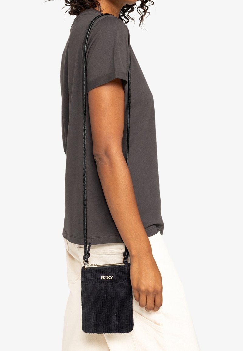 Personne portant une chemise à manches courtes gris foncé et un pantalon blanc, portant un petit sac bandoulière noir en velours côtelé avec le logo "Roxy".