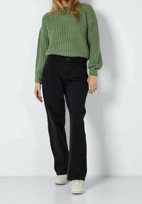 Grüner Strickpullover mit strukturiertem Muster, kombiniert mit schwarzen, weit geschnittenen Hosen und weißen Turnschuhen. Der Fokus liegt auf dem Material, der Schnitt ist locker.