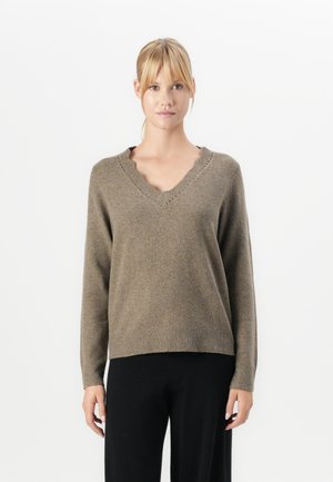Frau mit einem braunen V-Ausschnitt-Pullover mit gewellten Kanten und schwarzen Hosen, die vor einem schlichten weißen Hintergrund steht.