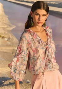 Floral-patterned sheer blouse in soft pink with ruffled neckline, long puffed sleeves, and a tied front. Worn with a light pink skirt.