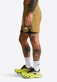 Men's khaki shorts with a black inner lining, elastic waistband, and logo; paired with neon yellow running shoes and white socks.