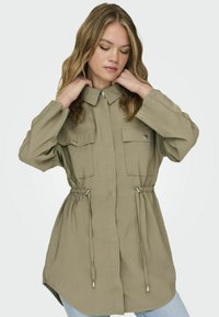 Khaki košeľová bunda s gombíkovým zapínaním, dvoma náprsnými vreckami, stiahnutým pásom a dlhými rukávmi. Mäkká tkanina, uvoľnený strih, zakrivený lem.