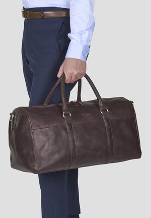 Brown leather duffle bag with a textured finish, dual top handles, and silver hardware, worn by a person in formal attire.