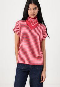 T-shirt rayé rouge et blanc avec un col en V et des manches retroussées, accompagné d'un bandana rouge à motifs autour du cou, porté avec un jean bleu.