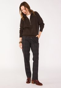 Sweatshirt zippé marron, chemise intérieure claire, pantalon à carreaux dans des tons foncés, bottines en cuir marron avec un talon épais et un bracelet.