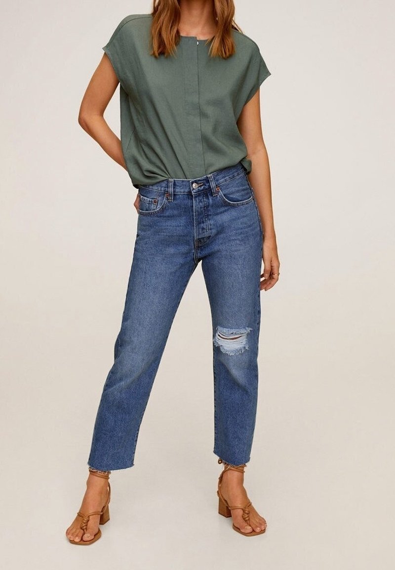 Femme portant un haut vert à manches courtes, un jean bleu taille haute avec déchirure au genou, et des sandales beige à brides avec talons blocs, debout sur un fond neutre.