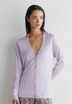 Kvinde med glat mørkt hår iført en lys lilla cardigan med knapper over mønstrede bukser, stående mod en ensfarvet hvid væg.