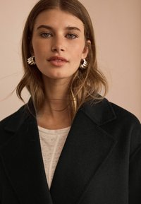 Svart ullkappa med slagkrage, bärs över en ljusfärgad topp. Silverörhängen med hoop-design ger detaljering. Bakgrunden är av en mjuk neutral ton.