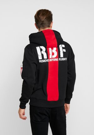 Mężczyzna ubrany w czarną bluzę z kapturem z pionowym czerwonym paskiem i białym napisem "RBF Remove Before Flight" na plecach, stojący na szarym tle.