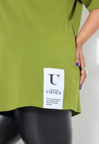 Camiseta de algodón verde de gran tamaño con una etiqueta lateral que presenta texto negro sobre tela blanca, combinada con leggins negros.