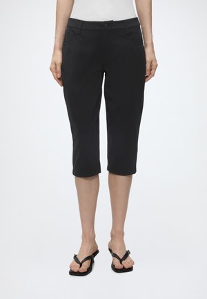Femme portant un pantalon noir au genou et des sandales noires, debout devant un fond blanc uni.