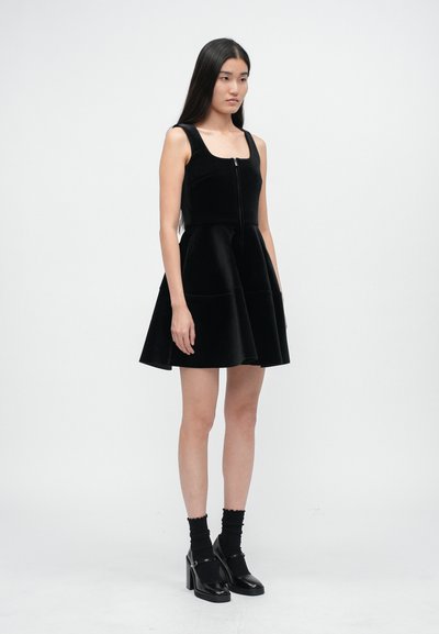 Femme debout portant une robe en velours noire sans manches avec une fermeture éclair à l'avant, des chaussettes noires et des chaussures Mary Jane à plateforme noires.