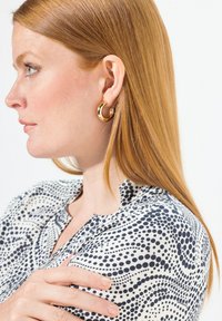 Goldene Creolen mit hochglanzpolierter Oberfläche. Das Modell trägt eine Bluse aus gemustertem Stoff mit marineblauen Punkten auf weißem Hintergrund.