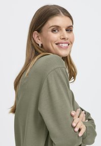 Top de manga larga verde oliva con un corte holgado, fabricado con un tejido suave y hombros caídos. La modelo tiene el pelo suelto y ondulado, y lleva pendientes de aro sencillos.
