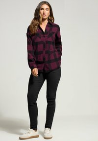 Vrouw in een paars en zwart geruit overhemd met knopen, zwarte skinny jeans en witte sneakers, staand met één hand in de zak.