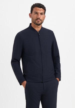 Man met donker krullend haar en baard, draagt een marineblauwe jas met rits en bijpassende broek, handen in de zakken, staand tegen een effen witte achtergrond.