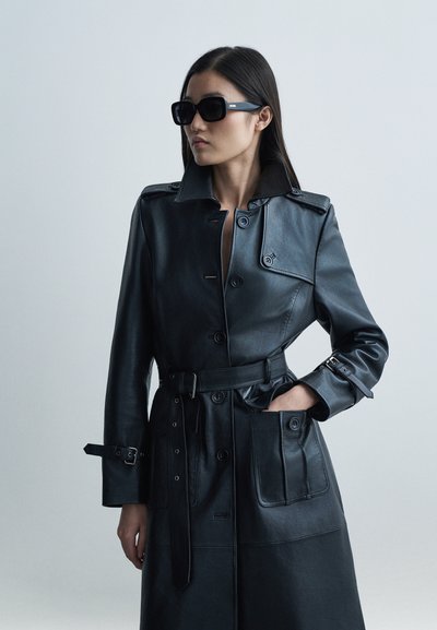 Črni usnjeni plašč z ovratnikom, pasom v predelu pasu, gumbi spredaj in stranskimi žepi. Gladek tekstura z rahlim sijajem.