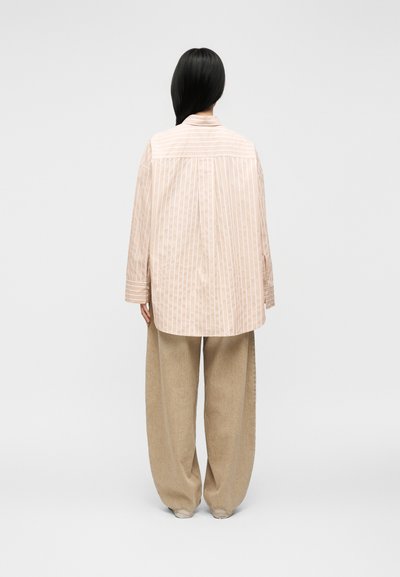 Femme aux longs cheveux noirs porte une chemise beige à rayures oversize et un pantalon large beige, debout de dos devant un fond uni.