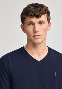 Granatowa koszulka typu v-neck wykonana z miękkiego materiału, z małym haftowanym logo po lewej stronie klatki piersiowej, gładka tekstura i prosty design.