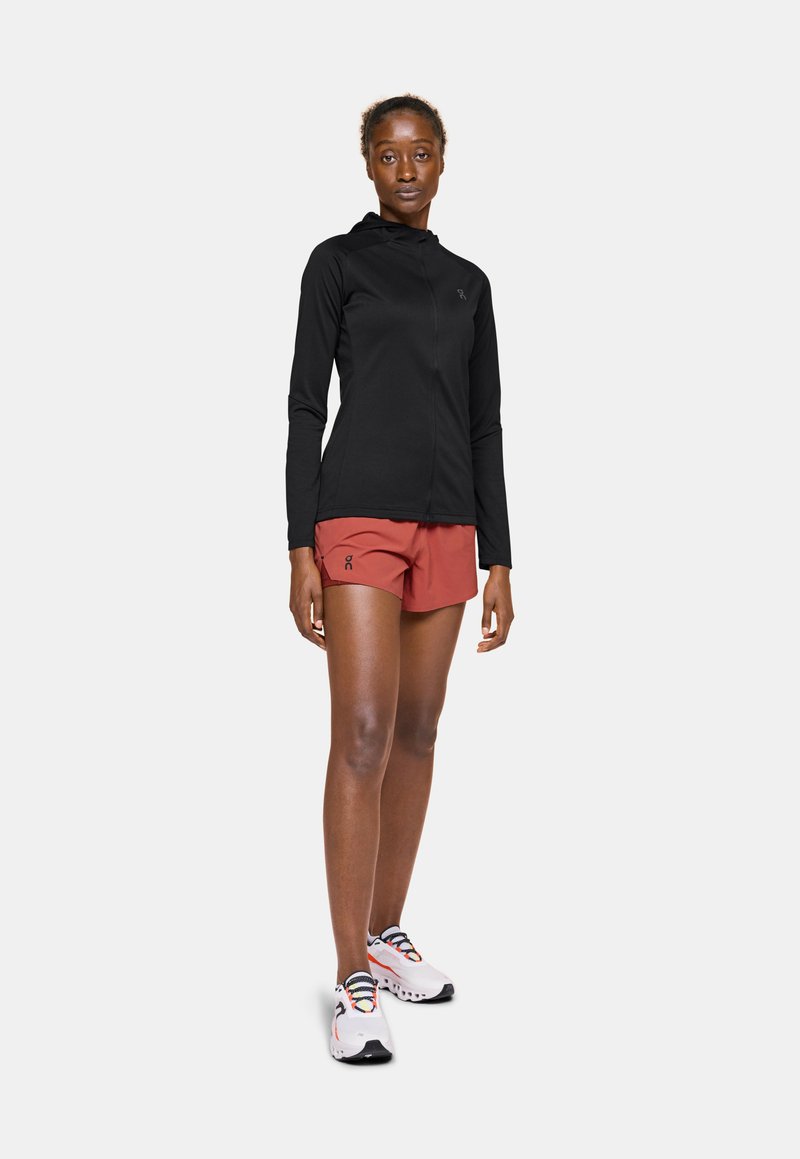 Schwarze, langärmelige Active-Jacke, rote Sportshorts und weiße Sneakers mit bunten Schnürsenkeln. Zeigt ein figurbetontes, aerodynamisches Design.