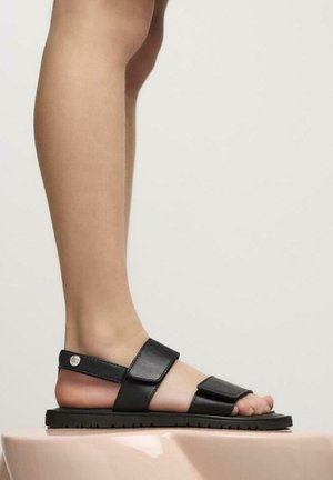 Sandale en cuir noir avec sangle réglable, semelle plate noire et design à bout ouvert. Texture lisse avec un accent matériel minimal.