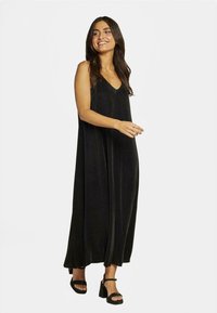 Robe maxi noire sans manches avec un décolleté en V, en tissu doux, présentant un design fluide et une texture subtile, associée à des sandales noires.