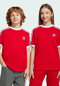 Röda idrottströjor med vita detaljer och Adidas-logotyp, kort ärm, rundad hals, mjukt bomullstyg, bärs med grå och röda byxor.