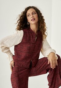 Chaleco de estampado rojo combinado con pantalones holgados, que presentan diseños florales, un cuello mao y botones decorativos. Tejido texturizado.