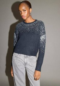 Maglione navy con decorazioni di paillettes sulle maniche, abbinato a jeans grigio chiaro con decorazioni sparse. Entrambi gli articoli hanno una vestibilità aderente.