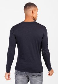 Man met kort haar en nek tatoeage, gekleed in een zwart shirt met lange mouwen en vervaagde grijze spijkerbroek, met de rug naar de camera gekeerd tegen een witte achtergrond.