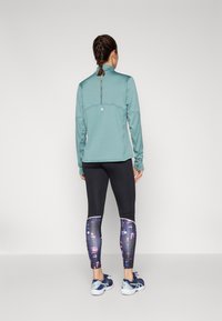 Haut à manches longues vert menthe avec un motif texturé, leggings noirs avec un imprimé abstrait coloré et chaussures de sport bleu marine.