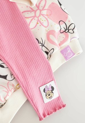 Manche rose côtelée avec un corps imprimé de fleurs et de personnages blancs, présentant une étiquette carrée avec le visage d'un personnage de dessin animé et des détails d'accent.