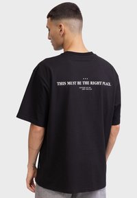 Zwart katoenen t-shirt met oversized ontwerp, korte mouwen en witte tekst op de achterkant met de woorden "DIT MOET DE JUISTE PLEK ZIJN."