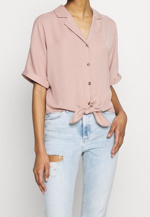 Woman wearing a short-sleeve pink button-up blouse tied at the waist, paired with light blue distressed jeans.