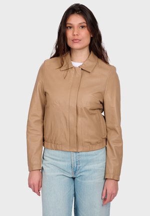 Junge Frau trägt eine hellbraune Lederjacke mit Kragen und hellblaue Jeans, steht vor einem schlichten, hellen Hintergrund.