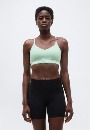 Fit person stående, iført lysegrøn Nike sports-bh og sorte shorts, med løve-tatovering på maven og navlepiercing.